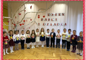 Dzieci elegancko ubrane na tle białego napisu i drzewka z sercami i napisem Dzień Babci i Dziadka.